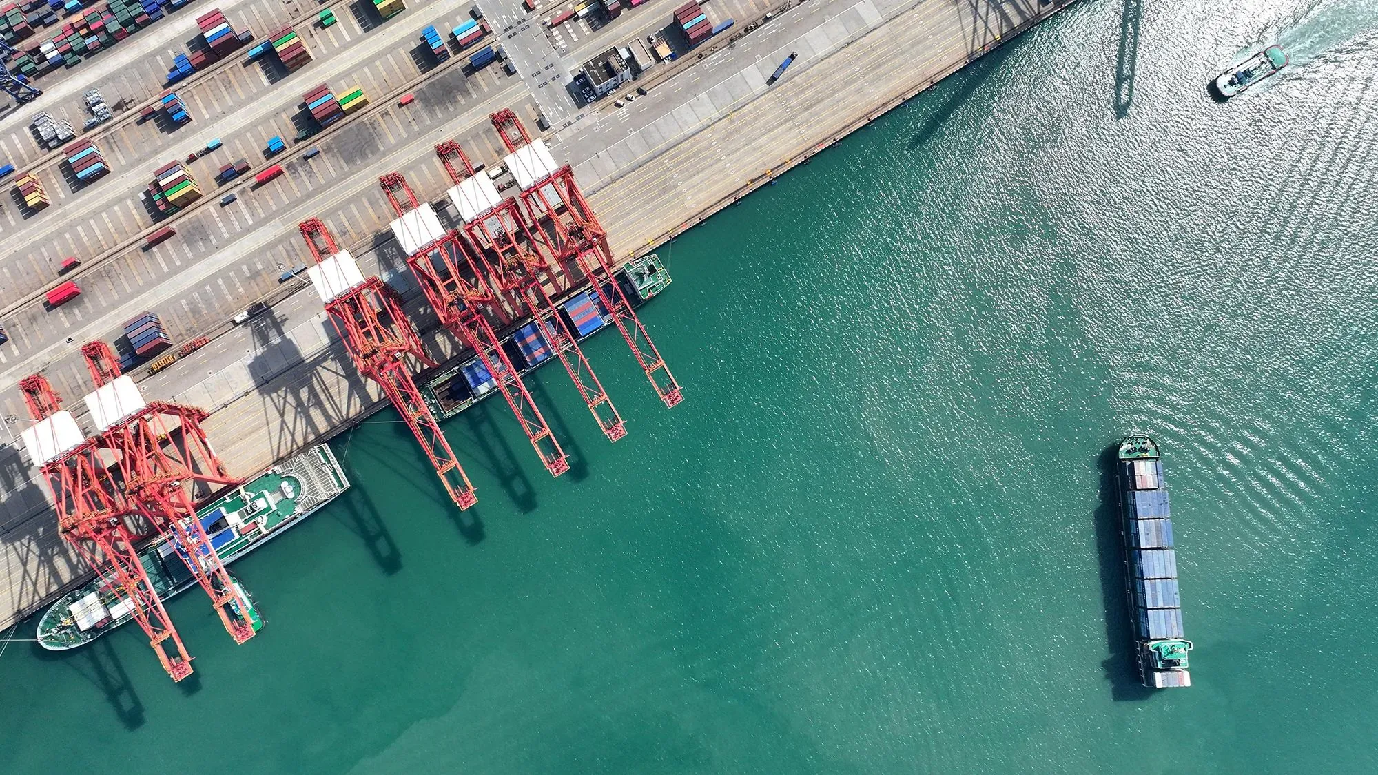 صورة جوية لميناء يحتوي على رافعات حمراء وحاويات شحن، مع سفينة تبحر في المياه القريبة، تعكس حركة التجارة الدولية.