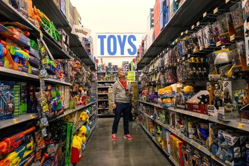 رجل يقف في ممر الألعاب داخل متجر، يتطلع إلى الرفوف المليئة بالألعاب المتنوعة، مع وجود علامة "ألعاب" بارزة في الأعلى.