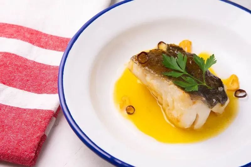 شرائح من سمك مطبوخ في صلصة زيت الزيتون، مزينة بأوراق البقدونس، تقدم في طبق أبيض مع حافة زرقاء، خلفية بيضاء مع منشفة حمراء.