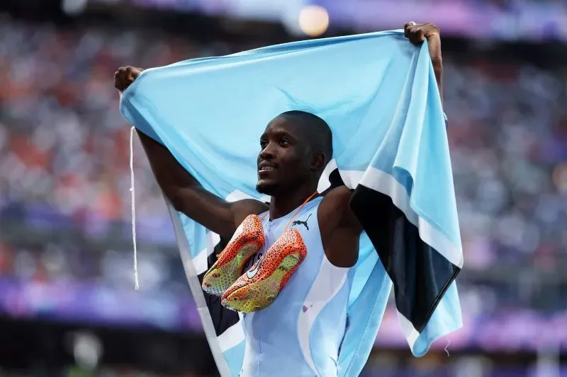 عداء يحمل علم بلاده ويظهر حذاءه الرياضي الملون بعد الفوز، يعكس طموح الشباب في رياضة ألعاب القوى.