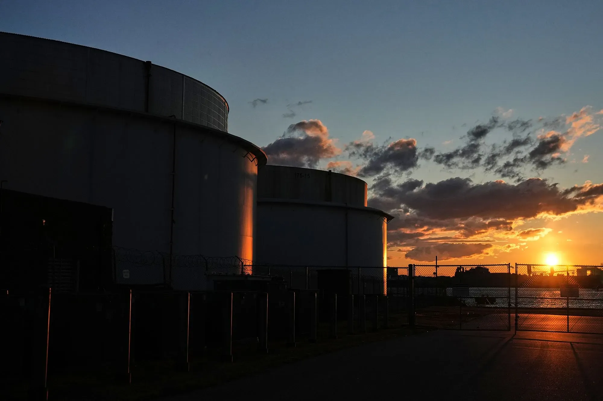 تظهر الصورة خزانات نفطية silhouetted ضد غروب الشمس، مما يرمز إلى التوترات في سوق النفط وتأثيرها على الأسعار العالمية.