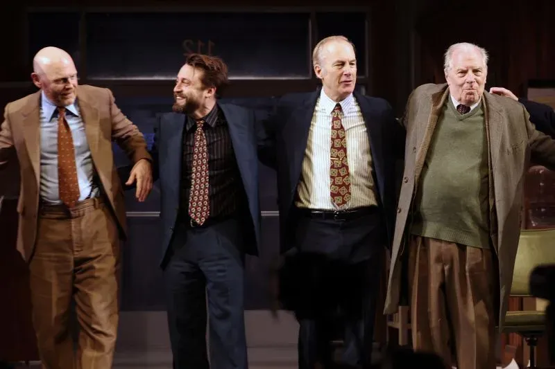 أربعة ممثلين يرتدون بدلات أنيقة، يقفون جنبًا إلى جنب على خشبة المسرح بعد انتهاء عرض مسرحي، مع تفاعل الجمهور في الخلفية.