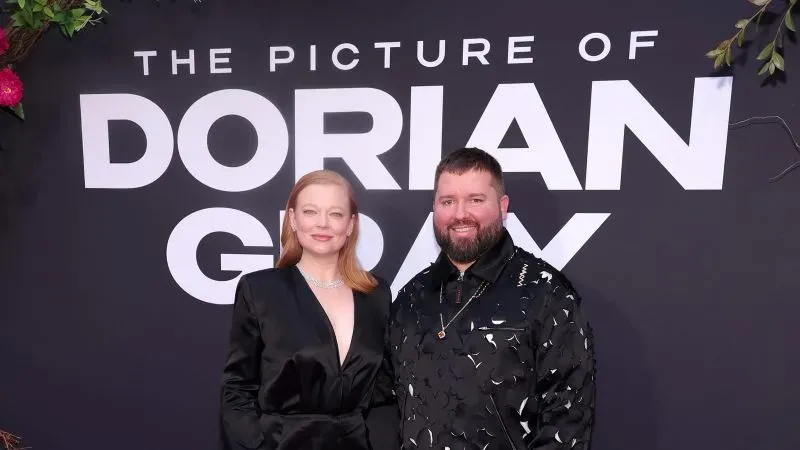 زوجان يقفان معًا أمام لافتة كبيرة تحمل عنوان "صورة دوريان غراي"، حيث يرتديان ملابس أنيقة تعكس أجواء المسرح.