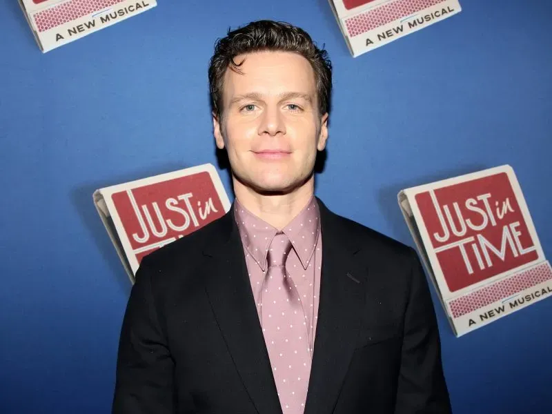 صورة لجوناثان جروف في حدث مسرحي، يرتدي بدلة أنيقة وقميص مزخرف، مع خلفية تحمل شعار "Just in Time".