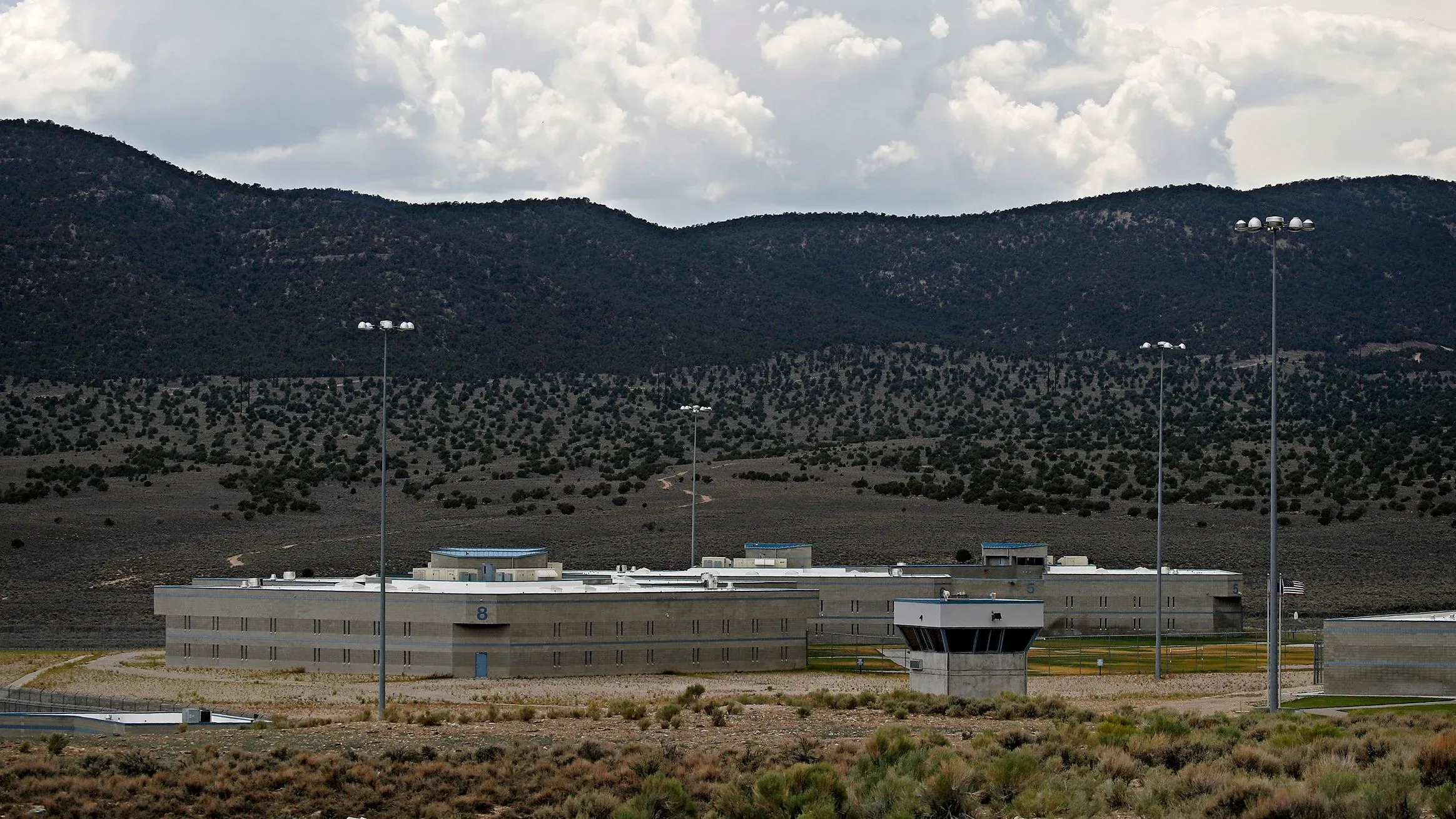 سجن ولاية إيلي في نيفادا، يظهر المبنى الرئيسي محاطًا بالجبال والسماء الملبدة بالغيوم، مع وجود أضواء عالية.