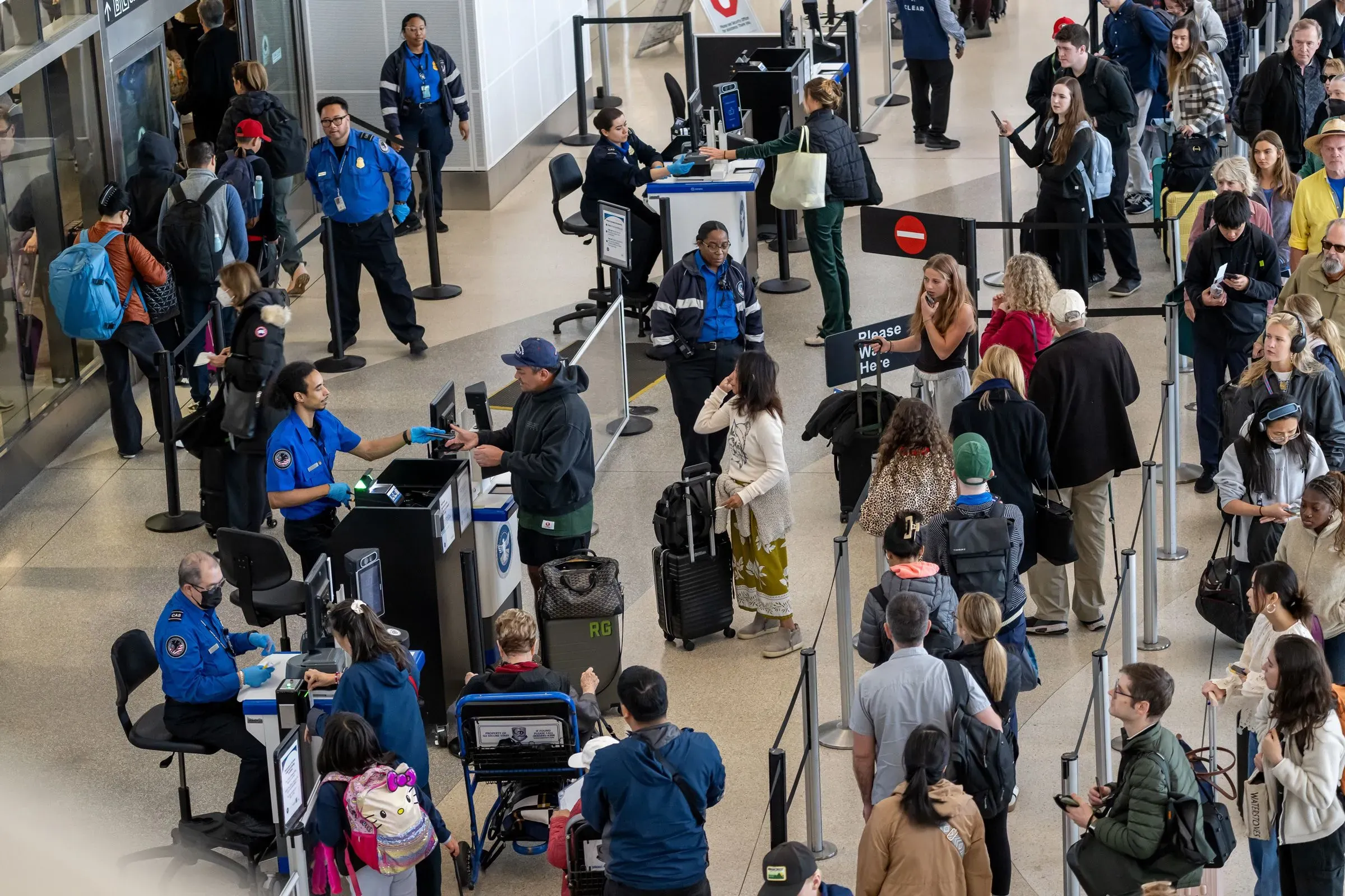 طوابير طويلة أمام نقاط التفتيش الأمنية في مطار مزدحم، مع موظفين من إدارة أمن النقل يتعاملون مع المسافرين.