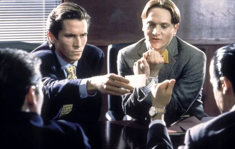 مشهد من فيلم "American Psycho" يظهر باتريك بيتمان وزملاءه في اجتماع عمل، يتبادلون بطاقات العمل في أجواء تنافسية.