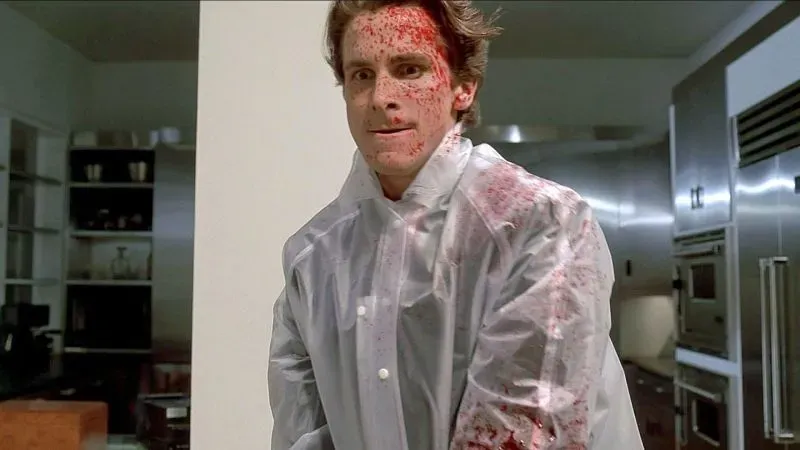 باتريك بيتمان، الشخصية الرئيسية في فيلم "American Psycho"، يرتدي معطفًا واقيًا مغطى بالدماء، ويظهر تعبيره القلق في مطبخ حديث.