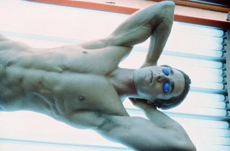 رجل عاري الصدر مستلقٍ تحت مصابيح الشمس الاصطناعية، يرتدي نظارات زرقاء للحماية، يعكس اهتمامه بروتين العناية بالبشرة.