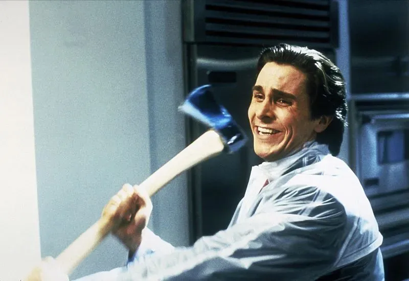 باتريك بيتمان مبتسمًا وهو يحمل فأسًا، يرتدي مئزرًا في مطبخ حديث، مما يعكس تناقض شخصيته بين الرغبة في الظهور الأنيق والجانب المظلم من حياته.