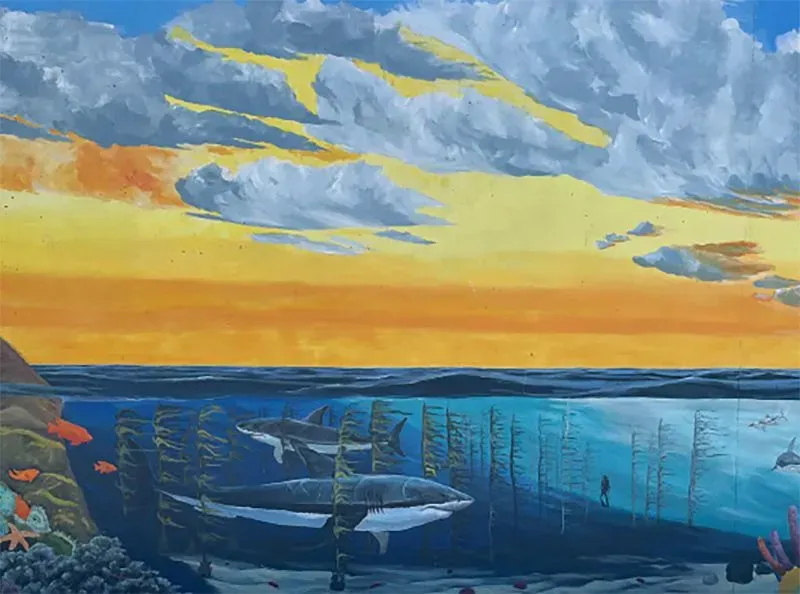 لوحة جدارية ملونة تُظهر مشهداً تحت الماء مع أسماك القرش وكائنات بحرية أخرى، تحت سماء غائمة تتلون بألوان الغروب.