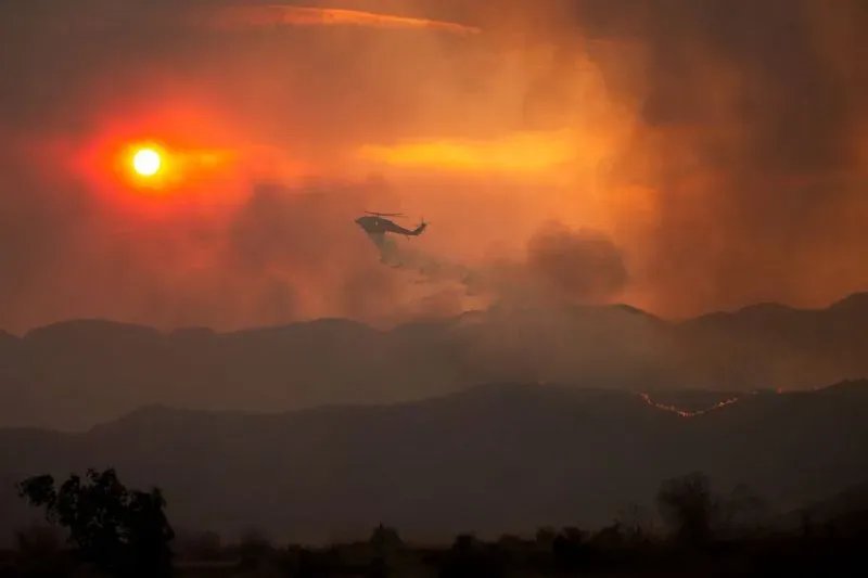 طائرة هليكوبتر تتصدى لحرائق الغابات في كاليفورنيا، مع ظهور الشمس خلف سحب الدخان، مشهد يعكس جهود الإطفاء تحت ظروف الجوية الخطيرة.