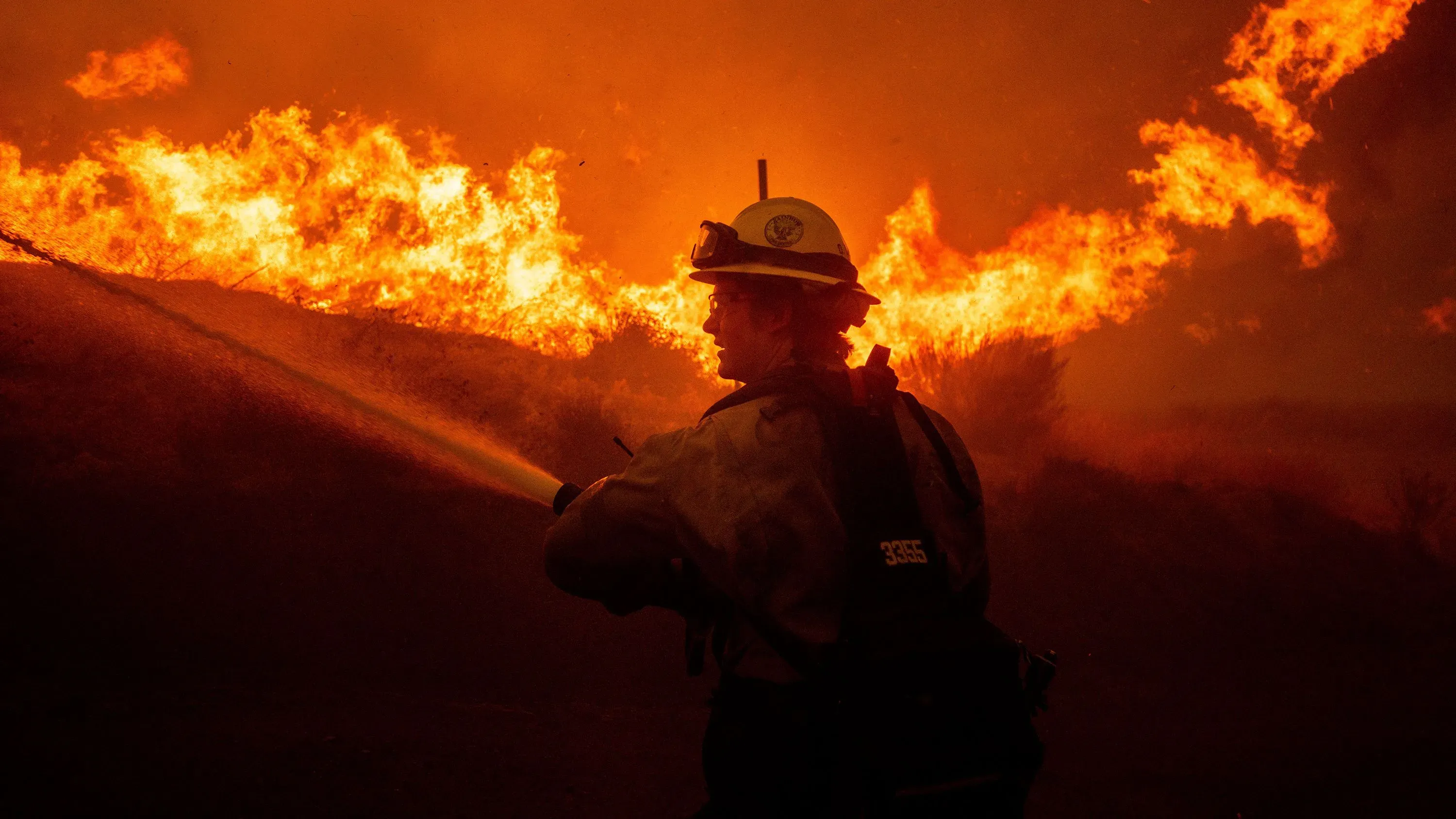 رجال إطفاء يكافحون حريقاً في كاليفورنيا، حيث تتصاعد النيران خلفهم بشكل مقلق، وسط ظروف جوية خطيرة.