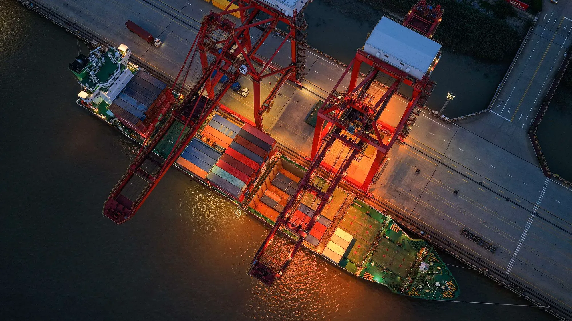 صورة جوية لميناء يحتوي على سفينة شحن محملة بالحاويات، مع رافعات حمراء تعمل على تفريغ الحمولات، تعكس حركة التجارة الدولية.