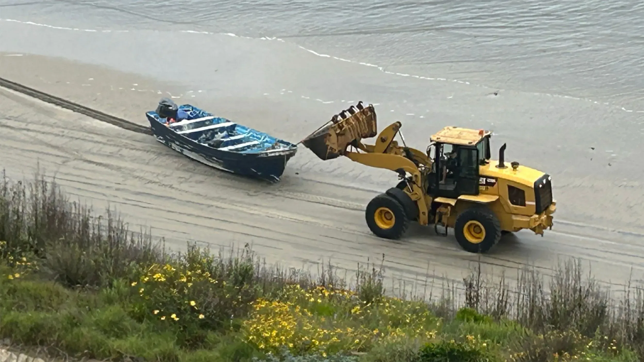 قارب صغير معطل على الشاطئ بينما يقوم جرافة برفعه، في سياق حادث انقلاب قارب قبالة سان دييغو.