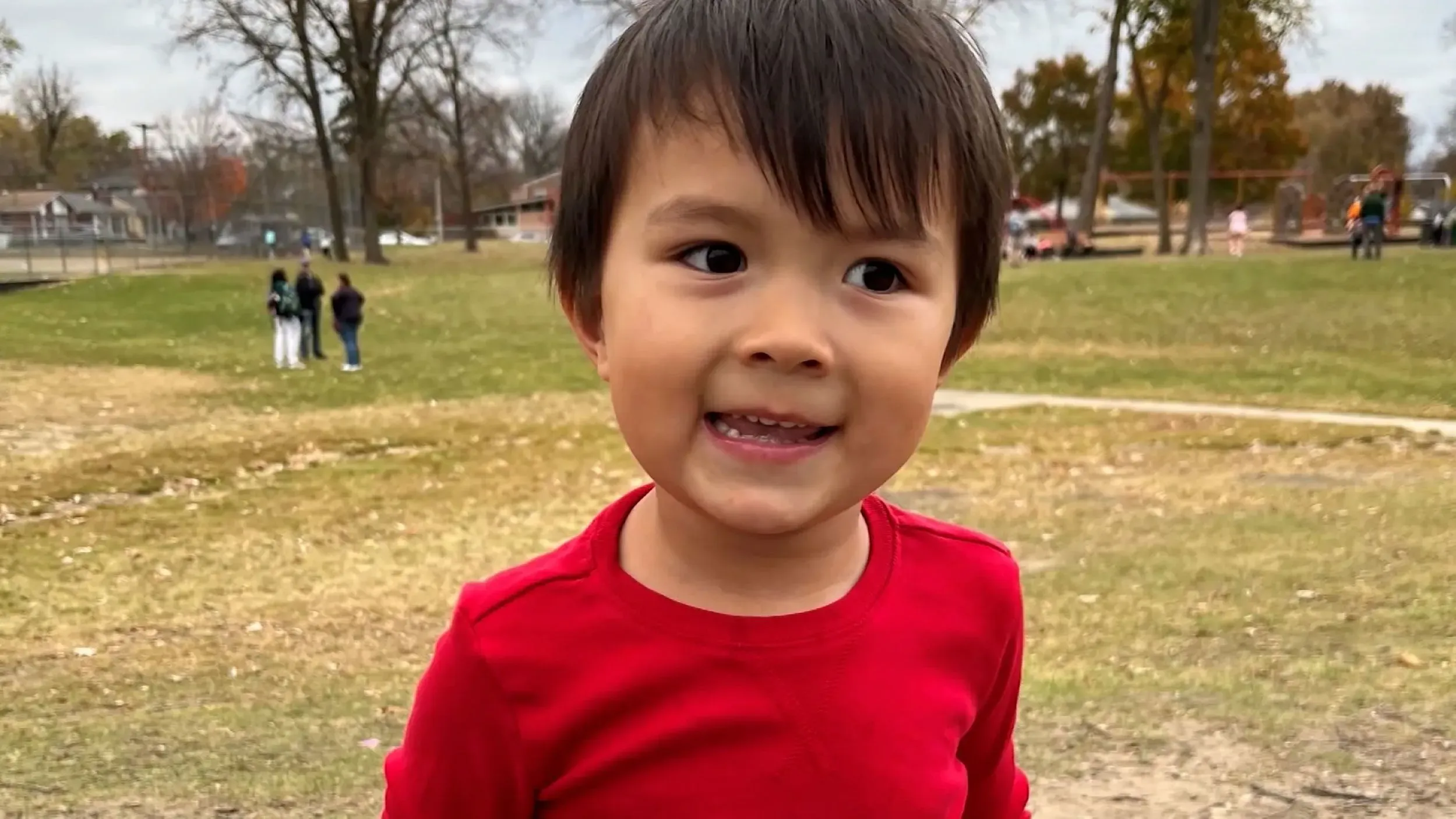 طفل صغير يرتدي قميصًا أحمر، مبتسمًا في حديقة، مع أشخاص آخرين في الخلفية، في سياق مأساة وفاة طفل في منشأة طبية.