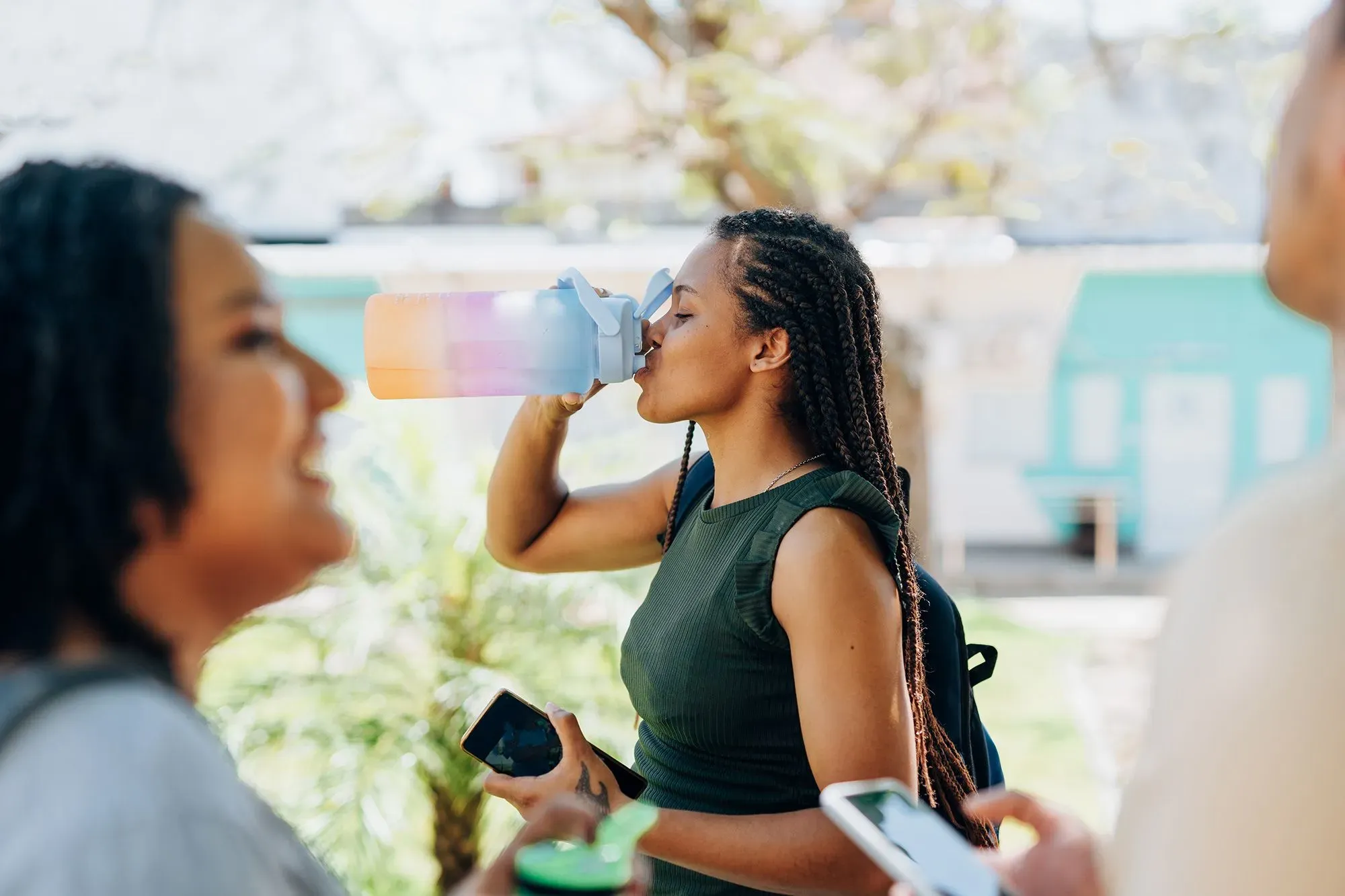 امرأة تشرب من زجاجة مياه قابلة لإعادة الاستخدام في حرم جامعي، محاطة بأصدقاء، مما يعكس أسلوب حياة مستدام واهتمامًا بالبيئة.