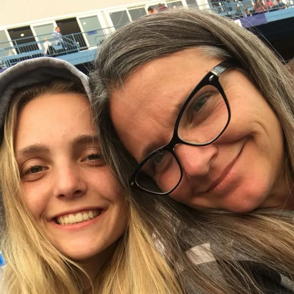 صورة لامرأة ذات شعر رمادي ترتدي نظارات، تبتسم مع فتاة شابة ذات شعر أشقر. تعكس الصورة علاقة عائلية ودعماً في ظل الظروف الصعبة.