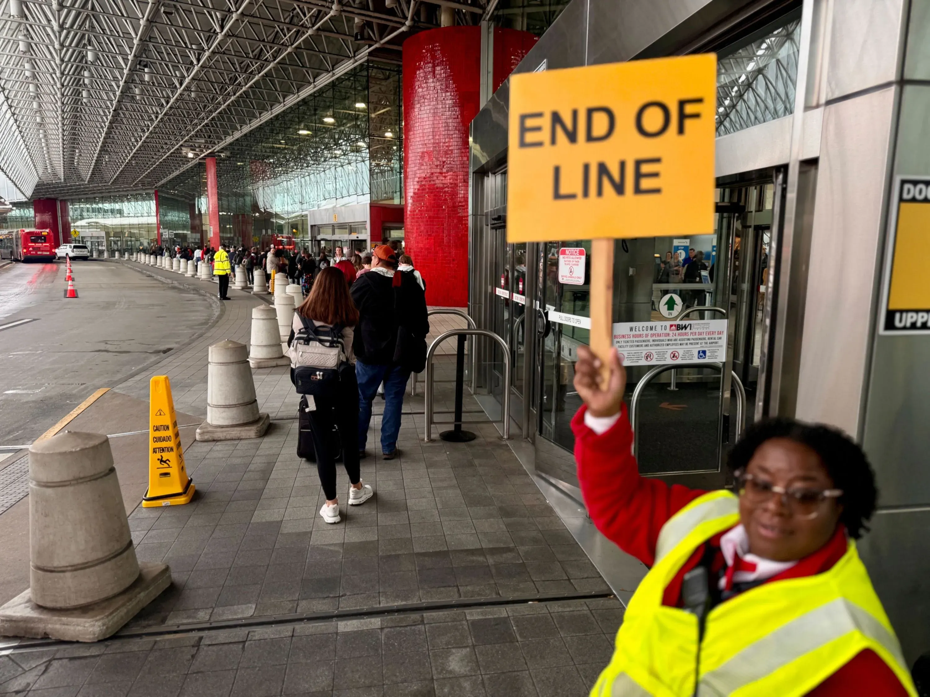 طوابير طويلة من المسافرين في مطار، مع موظف يحمل لافتة "نهاية الطابور"، تعكس تأثير نقص موظفي إدارة أمن النقل بسبب الإغلاق الحكومي.