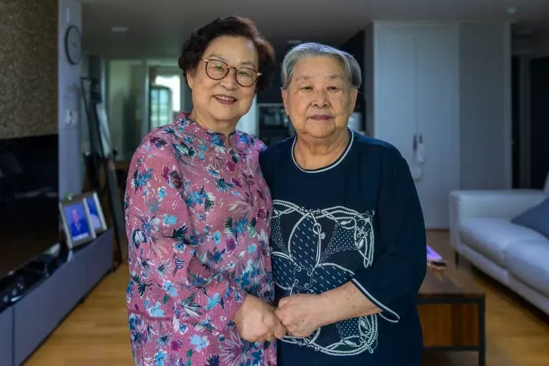 مين يونغ جاي وشقيقتها الكبرى تتعانقان في منزل حديث، تعكسان ذكريات عائلتهما السعيدة قبل الحرب الكورية.