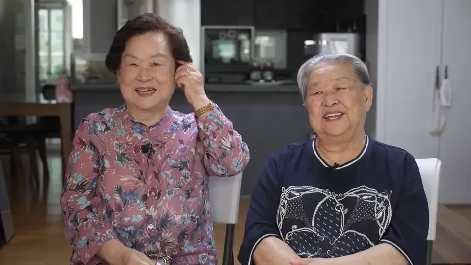 امرأتان مسنّتان تجلسان معًا، تبتسمان في غرفة معيشة حديثة، تعكسان ذكريات عائلتهما في زمن الحرب الكورية وأثرها.