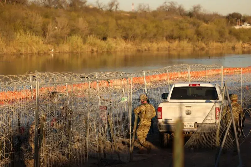 جنود يرتدون زيًا عسكريًا يعملون على الحدود الأمريكية مع وجود سياج من الأسلاك الشائكة وخلفهم سيارة بيك أب، مما يعكس التصعيد في جهود مراقبة الهجرة.
