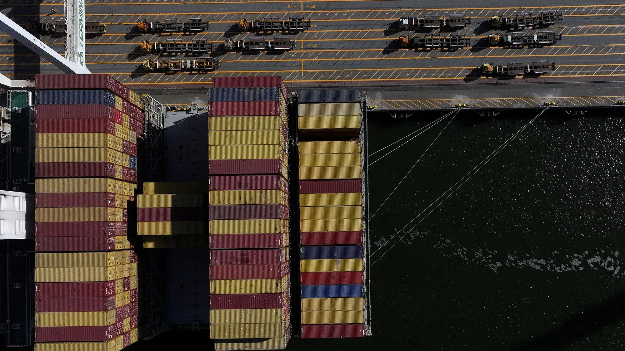 صورة توضح حاويات شحن ملونة في ميناء، مع شاحنات مخصصة لنقل البضائع، تعكس تأثير الحرب التجارية على الاقتصاد العالمي.