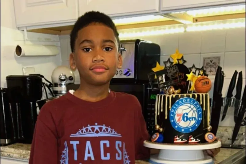 طفل يحمل كعكة عيد ميلاد مزينة بشعار فريق فيلادلفيا 76ers، يقف في مطبخ منزلي مع أدوات مطبخ خلفه.