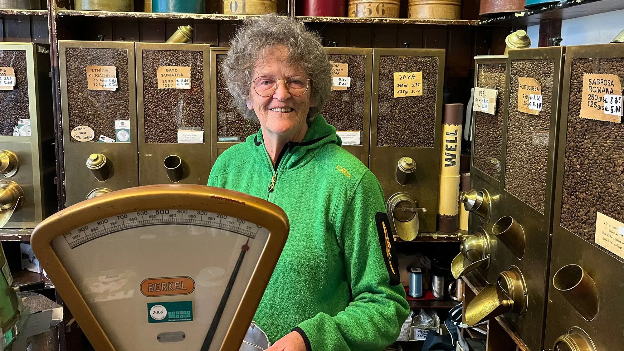 امرأة مسنّة ترتدي سترة خضراء تقف أمام أرفف مليئة بحبوب القهوة في متجر 't Zonnetje التاريخي بأمستردام، مع ميزان بيركل قديم.