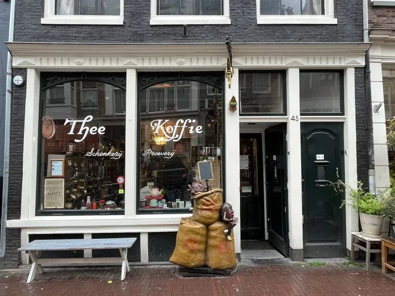 واجهة متجر "t Zonnetje" في أمستردام، مع لافتة مذهبة وشخصيات مرحة، تعكس تاريخًا عريقًا في تقديم الشاي والقهوة منذ القرن السابع عشر.