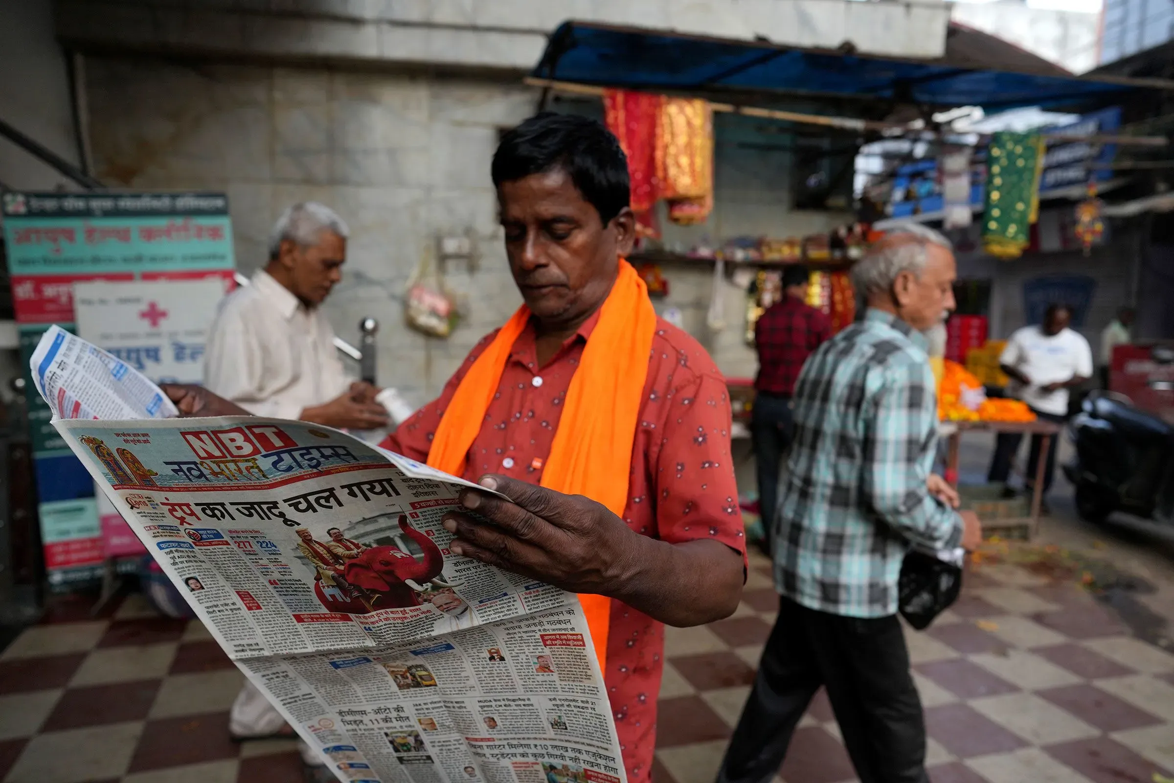رجل يرتدي سترة برتقالية يقرأ صحيفة في سوق مزدحم، مع وجود أشخاص آخرين في الخلفية، مما يعكس التفاعل اليومي مع وسائل الإعلام في الهند.