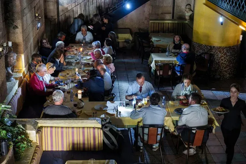 مجموعة من الناس يجلسون حول طاولات في مطعم مضاء بشموع، يتناولون الطعام في أجواء هادئة أثناء انقطاع الكهرباء في إسبانيا.