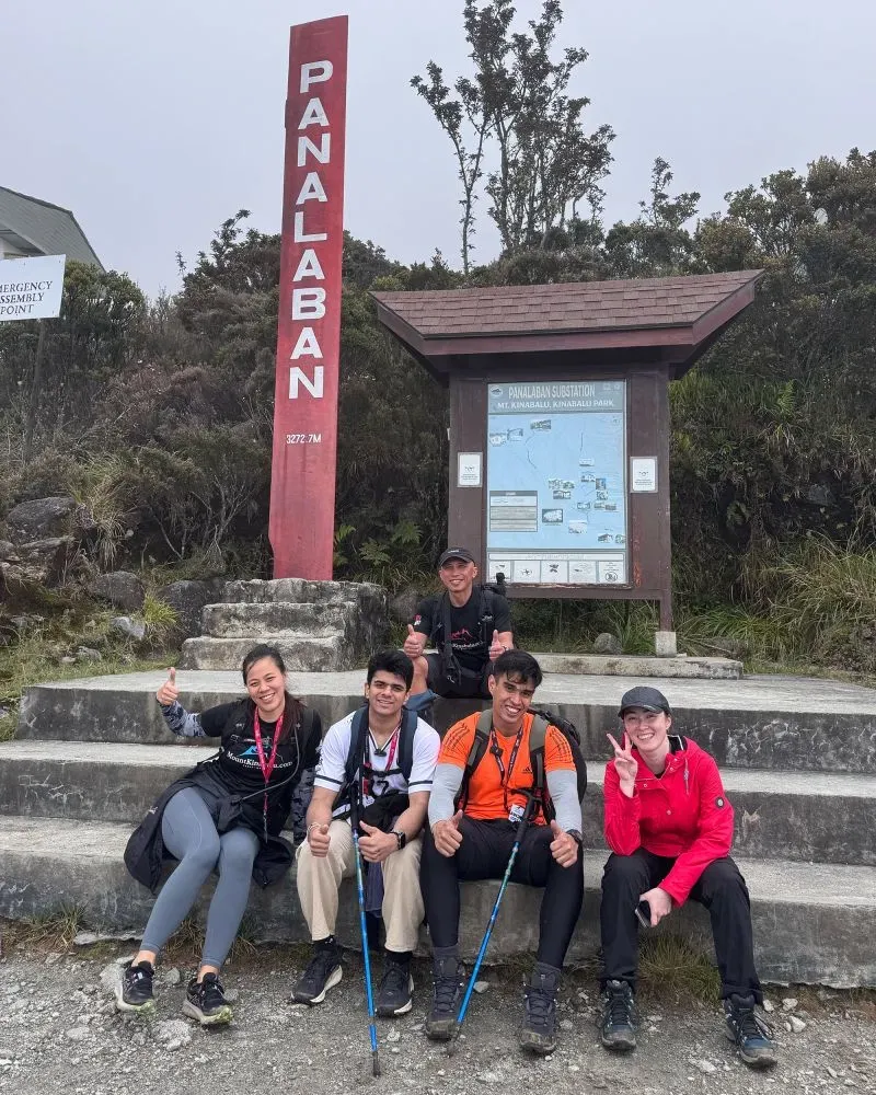 مجموعة من خمسة متسلقين يجلسون على درجات صخرية، مبتسمين ويقدمون إيماءات إيجابية، مع لافتة تشير إلى نقطة تجميع الطوارئ في جبل كينابالو.