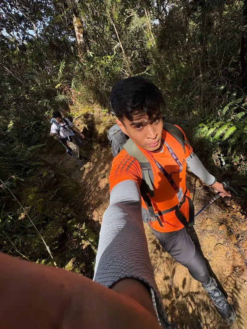 شخصان يتسلقان جبل كينابالو، يرتدي أحدهما قميصاً برتقالياً زاهياً، بينما يظهر الآخر في الخلفية. تتخلل المناظر الطبيعية الخضراء المحيطة.