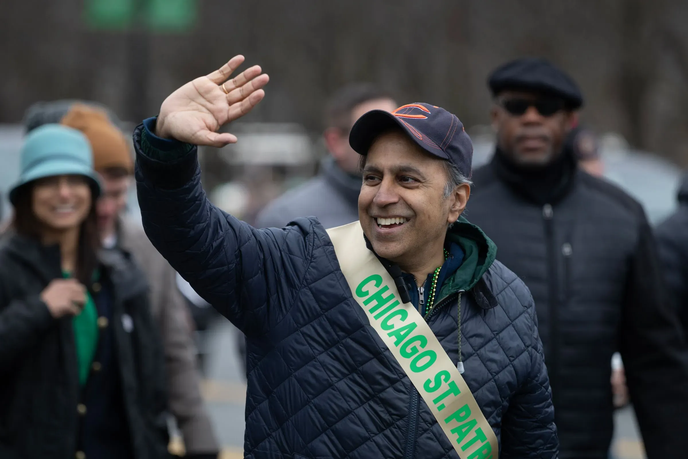 رجل مبتسم يرتدي سترة داكنة وقبعة، يلوح بيده في احتفال يوم القديس باتريك في شيكاغو، مع وجود حشود في الخلفية.