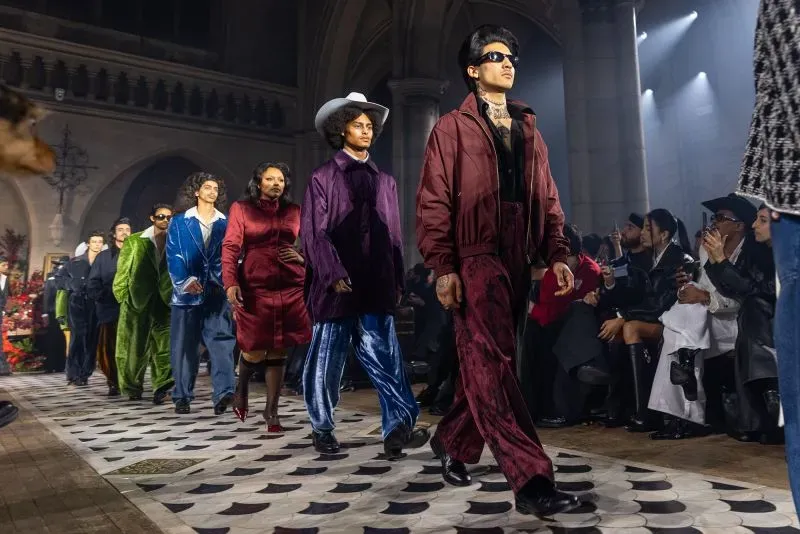 عرض أزياء يضم مجموعة من الرجال يرتدون ملابس ملونة وأنيقة، يتقدمون على منصة عرض في مكان فني مع جمهور يتابعهم.