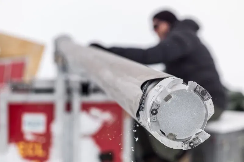 أنبوب حفر عمودي يستخدم لجمع عينات من الجليد في غرينلاند، مع شخص يعمل في الخلفية. يساهم في الأبحاث المناخية الهامة.