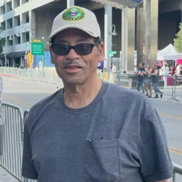 رجل يرتدي نظارات شمسية وقبعة، يقف في منطقة حضرية، مع وجود حواجز في الخلفية، يعكس تأثير الحرائق المدمرة في لوس أنجلوس.