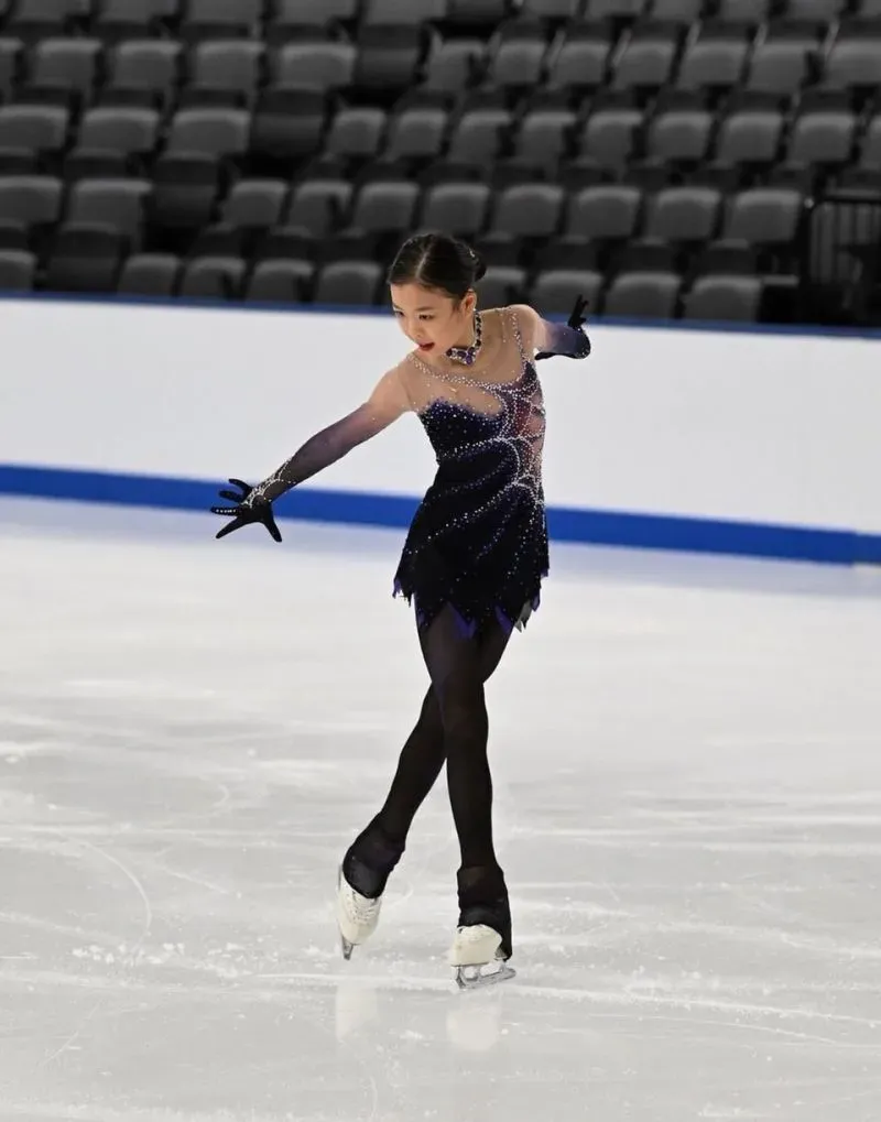 لاعبة تتزلج على الجليد ترتدي فستانًا أسود مزخرفًا بالأحجار اللامعة، تؤدي حركات أنيقة في ساحة خالية من الجمهور.