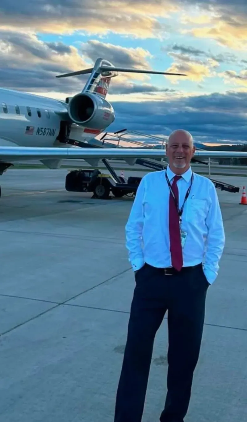 صورة لرجل يرتدي زياً رسمياً يقف أمام طائرة صغيرة على مدرج، حيث تشرق الشمس في السماء. يظهر الحزن في هذه الصورة بعد حادثة التصادم المأساوية.