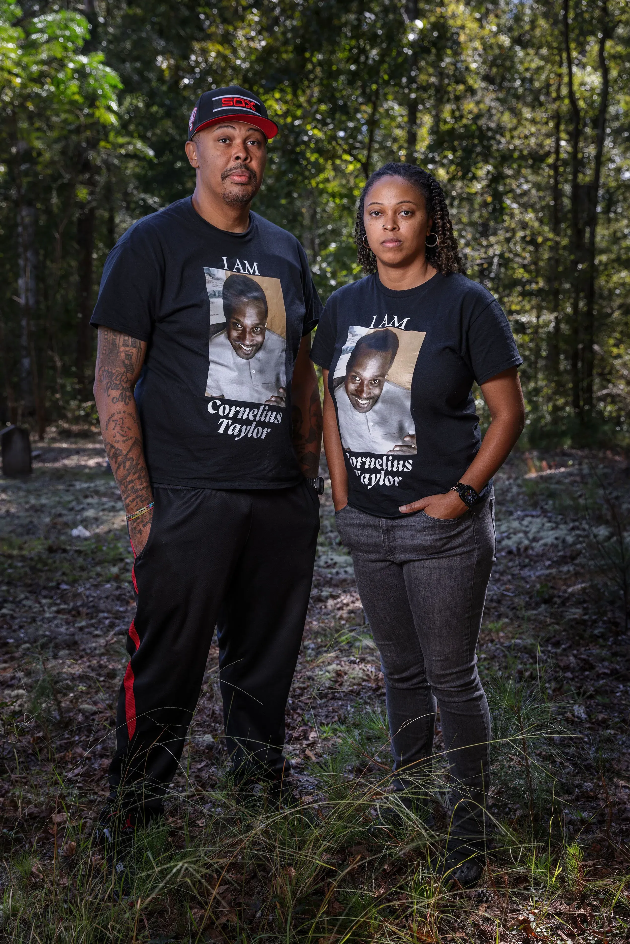 زوجان يرتديان قمصان مكتوب عليها "أنا كورنيليوس تايلور" يقفان في غابة، مع تعبيرات جدية تعكس التأثر بقصة حياة كورنيليوس.