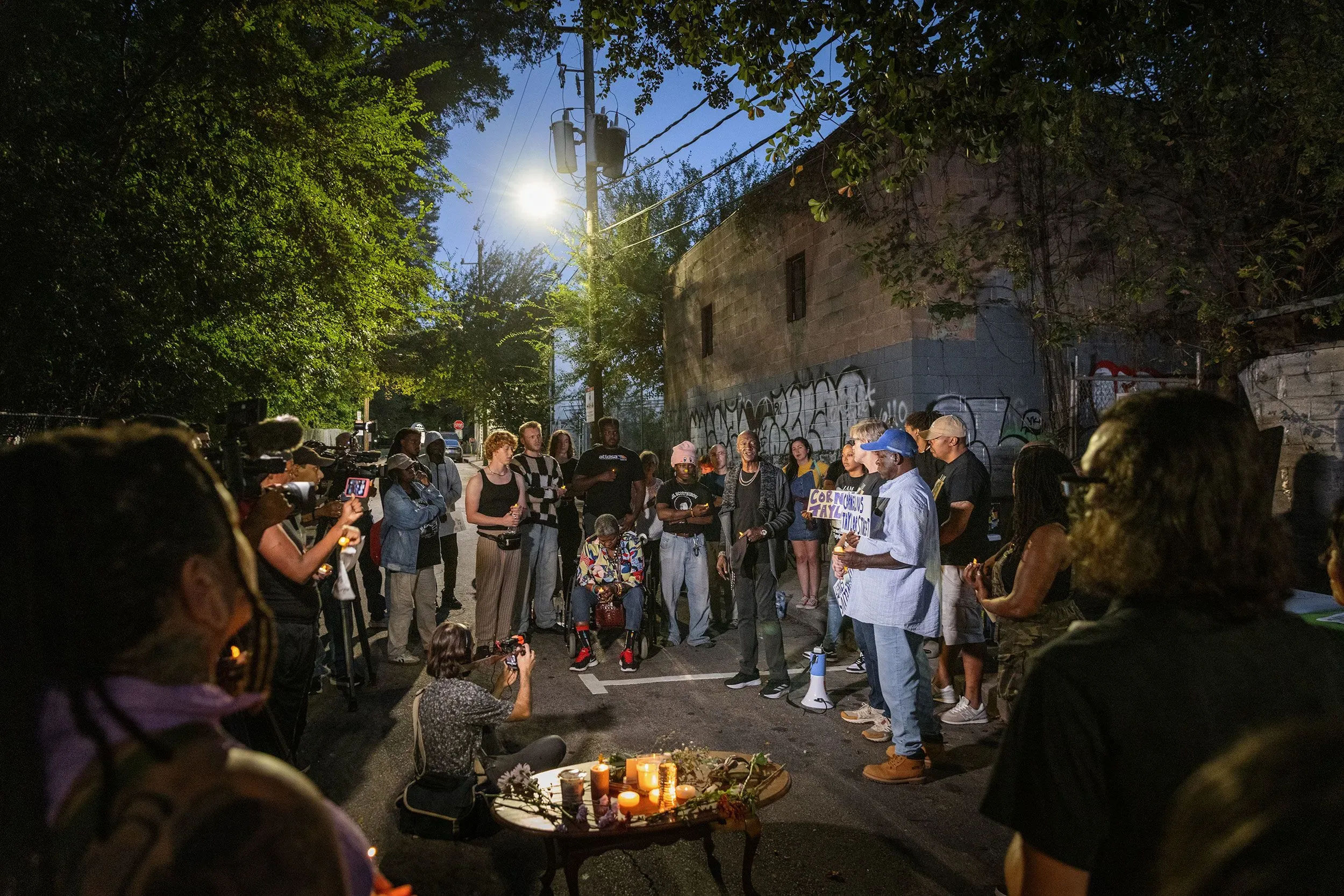 تجمع حشد من الأشخاص في منطقة مظلمة بأتلانتا لإحياء ذكرى كورنيليوس تايلور، مع وجود شموع وأعلام، تعبيرًا عن الحزن بسبب وفاته المأساوية.