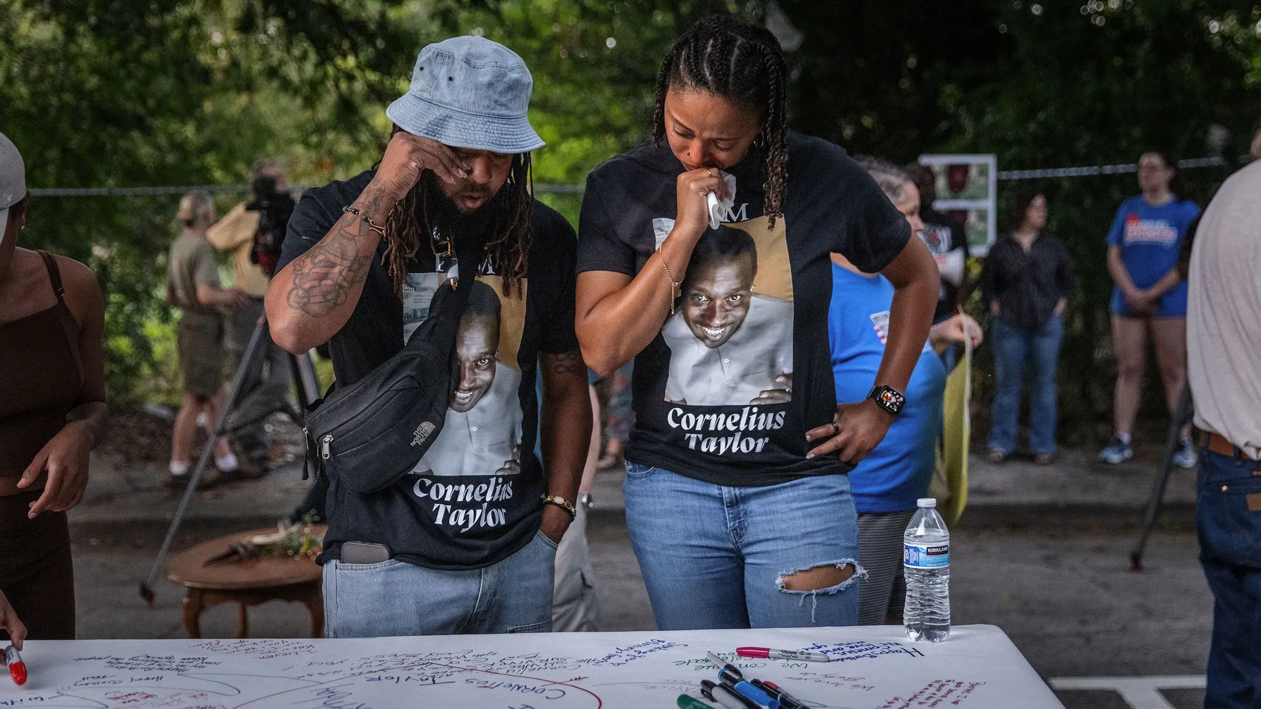 تظهر الصورة مجموعة من الأشخاص يرتدون قمصان تحمل صورة كورنيليوس تايلور، وهم يتذكرون حياته في حدث تأبيني، مع مشاعر الحزن والتأمل.