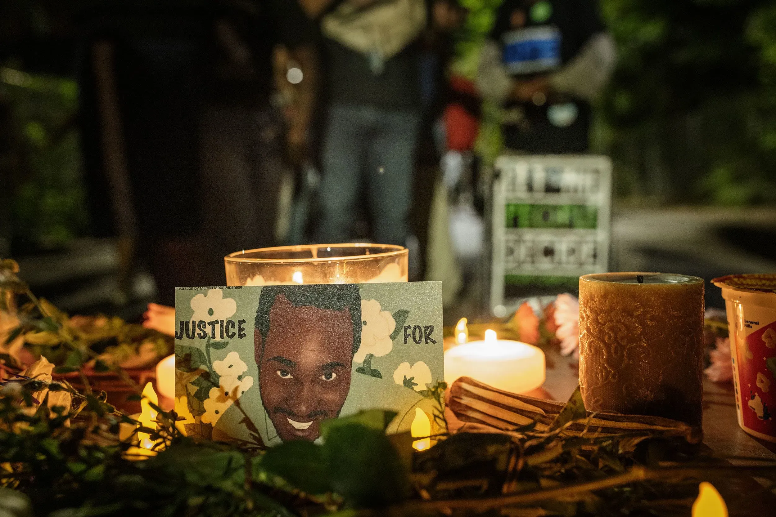 شمعة مضاءة وصورة لكورنيليوس تايلور محاطة بأزهار وعبوات، تعبيرًا عن الحزن والمطالبة بالعدالة بعد وفاته المأساوية أثناء إزالة مخيم المشردين.