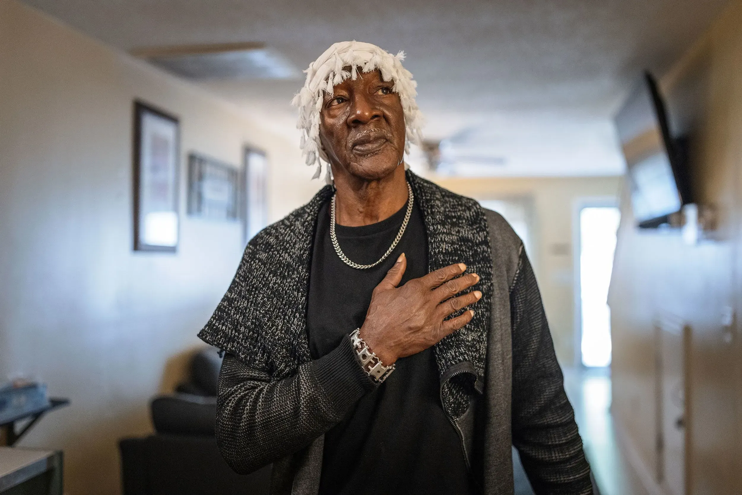 كورنيليوس تايلور، رجل مسن يرتدي سترة داكنة، يقف في ممر داخل مبنى، مع تعبير جاد، يعكس التحديات التي واجهها في حياته.