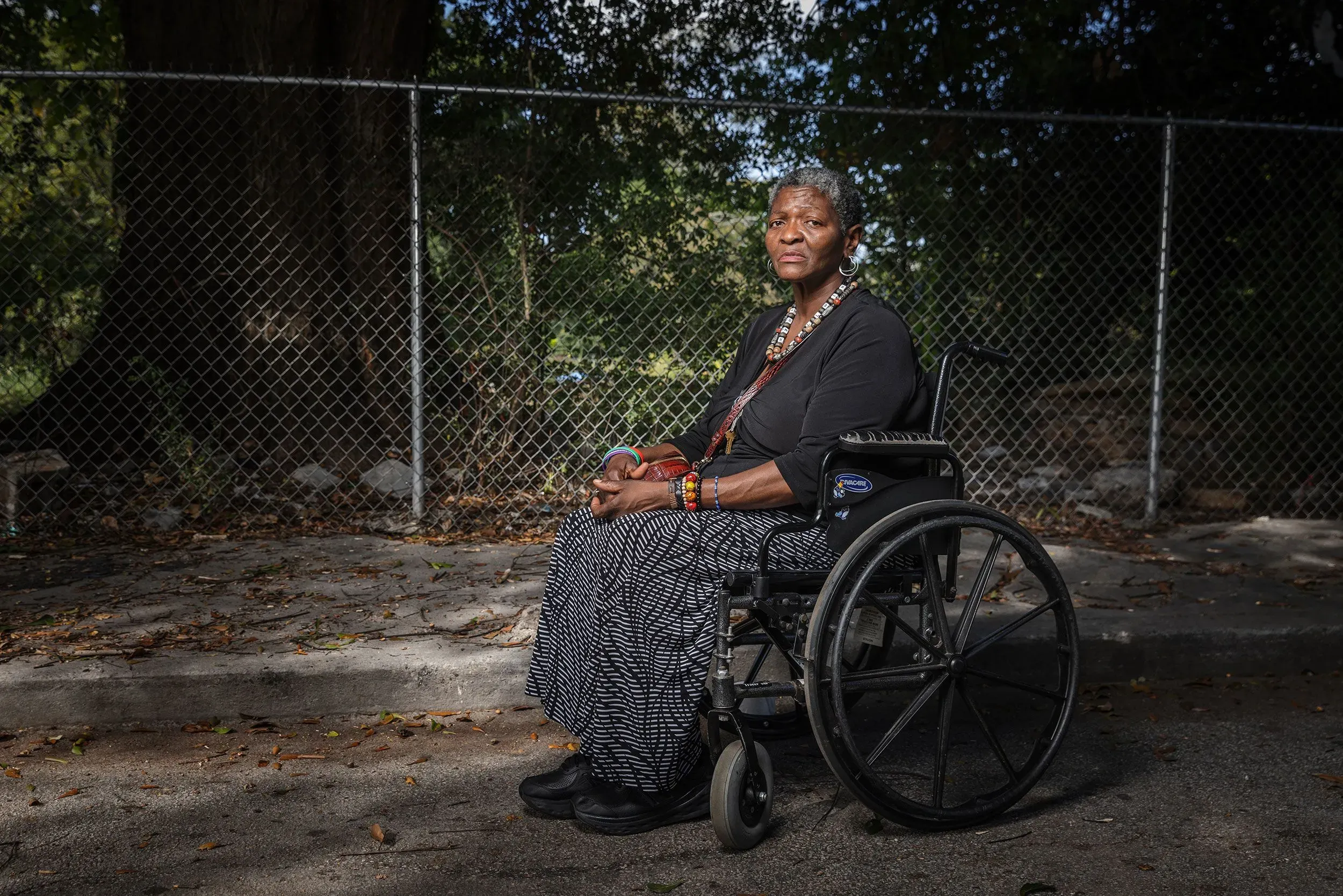 امرأة جالسة في كرسي متحرك، ترتدي ملابس تقليدية، وتظهر ملامح القوة والتحدي، بينما تتواجد في منطقة خارجية محاطة بسياج.