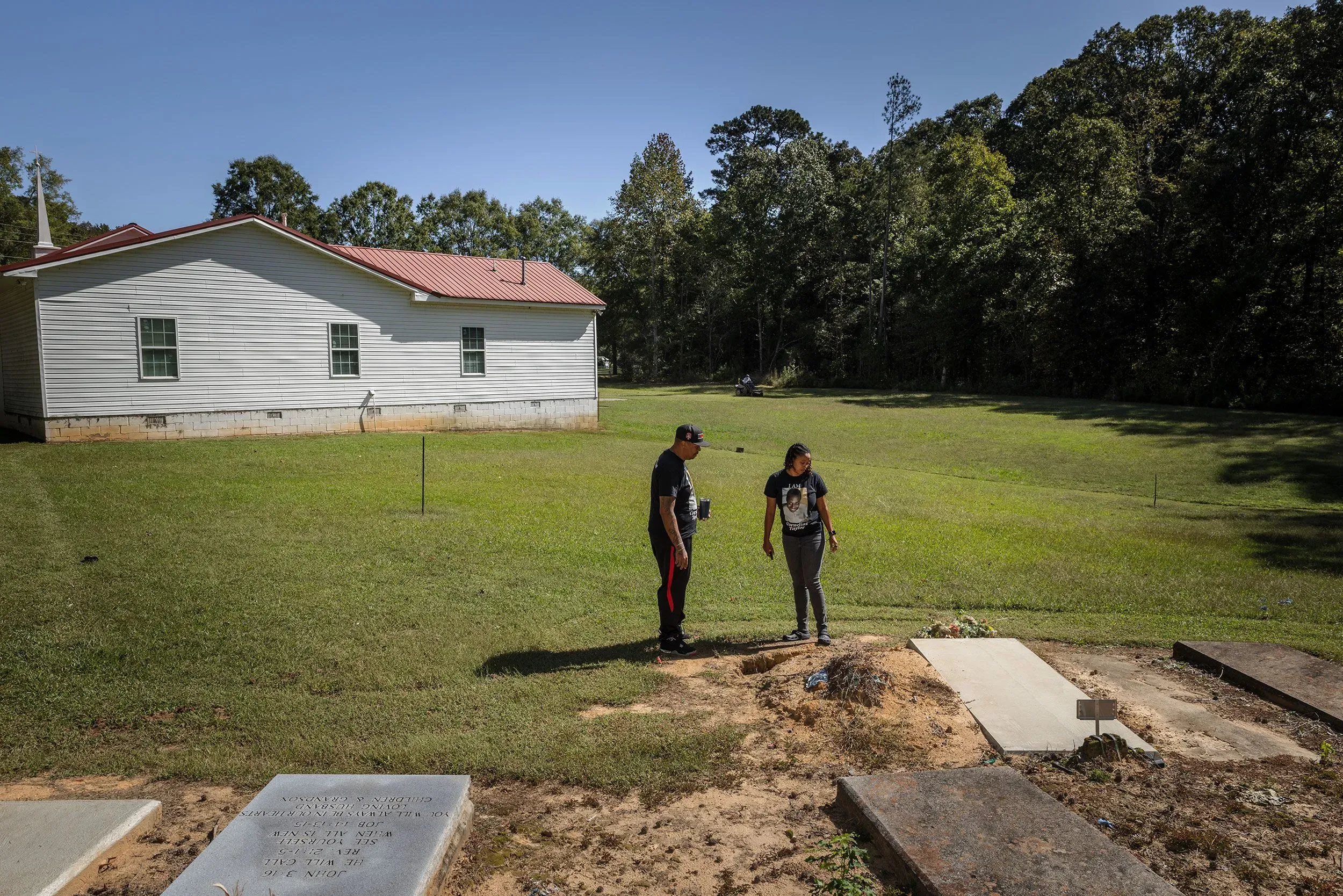 شخصان يقفان أمام قبر في منطقة خضراء، مع منزل أبيض في الخلفية، يعكسان لحظة تأمل وحزن على فقدان كورنيليوس تايلور.