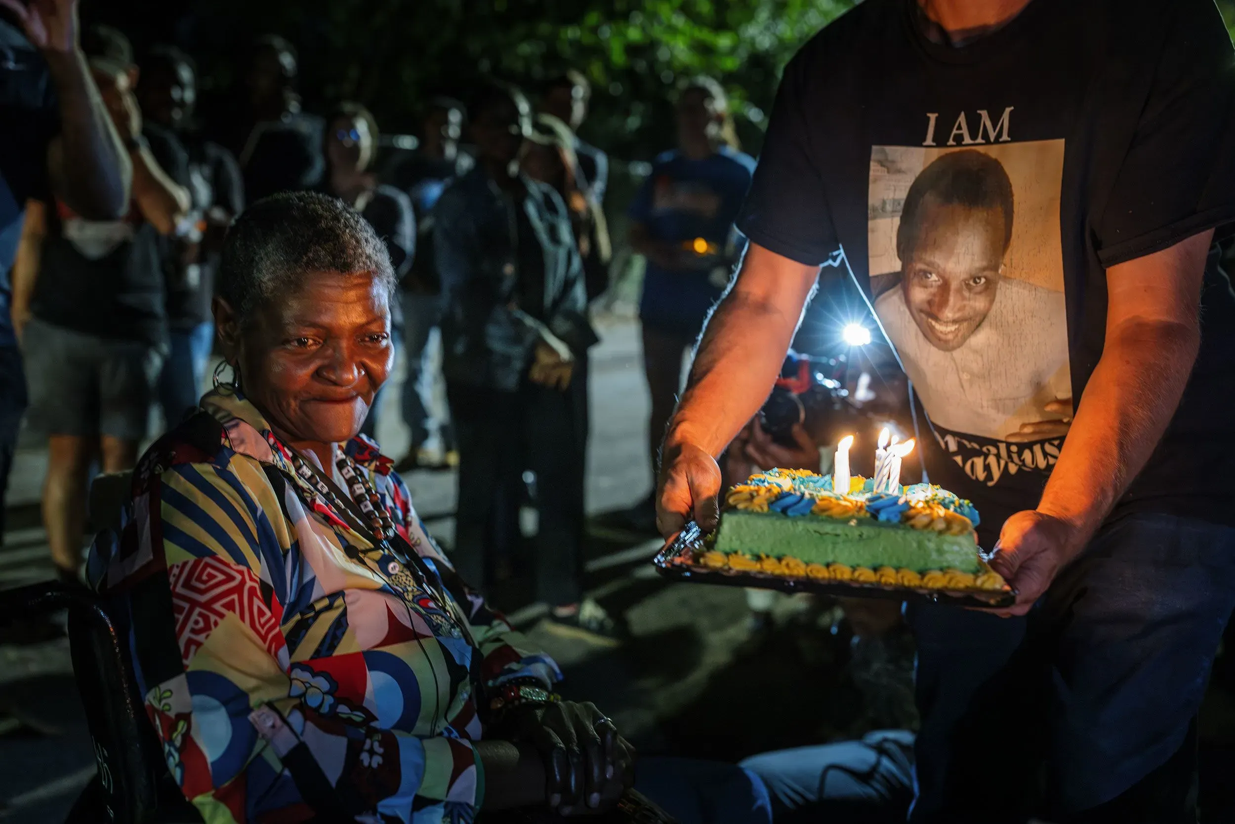 امرأة مسنّة مبتسمة تجلس على كرسي متحرك، بينما يحمل شخص آخر كعكة مزينة بالشمع. خلفهم، تجمع من الناس في أمسية احتفالية.