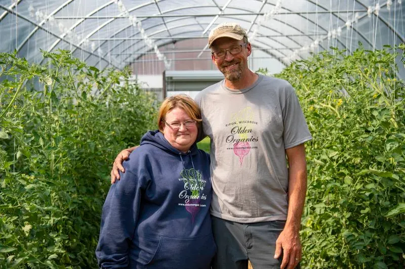 زوجان يقفان معًا في دفيئة مليئة بالنباتات الخضراء، يمثلان المزارعين المحليين في ويسكونسن الذين يواجهون تحديات التمويل.