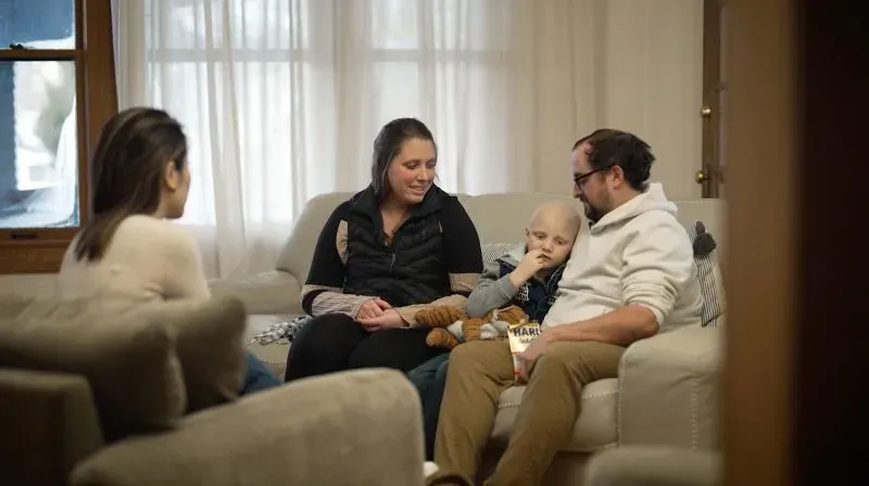 عائلة فيلا تجلس مع كايلين، الطفل المصاب بالسرطان، في غرفة معيشة مريحة، تعكس تحدياتهم اليومية في مواجهة المرض.